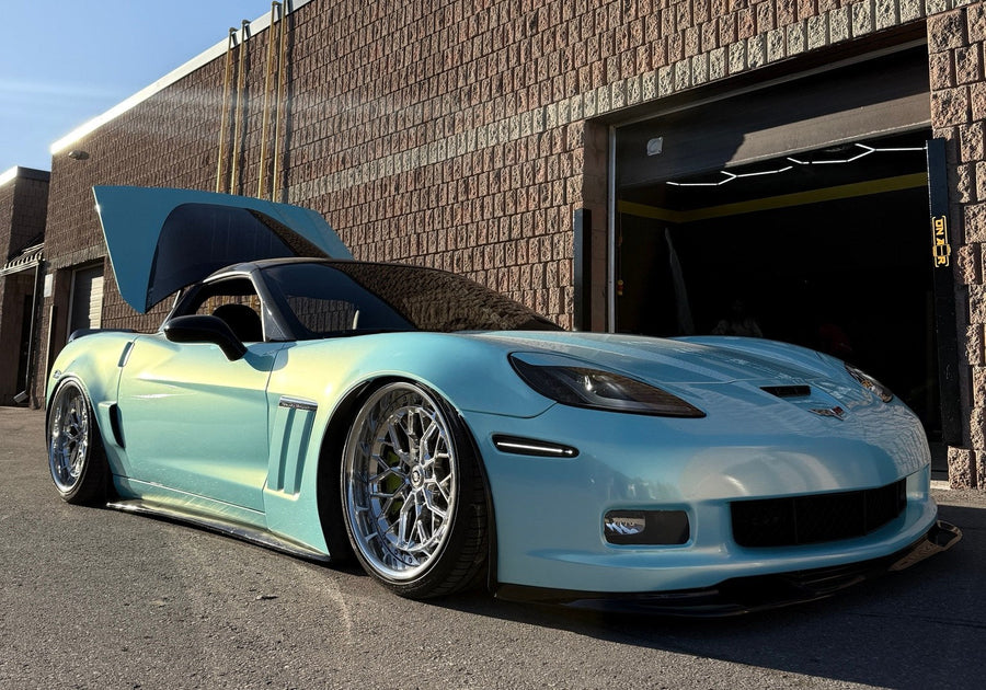 Azad's turquoise bagged C6 Corvette aired out outside ONAIR, with polished wheels, a black splitter and its rear hatch open.