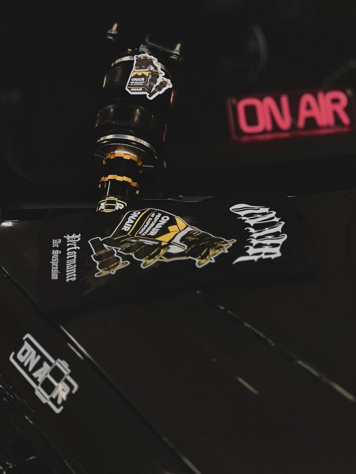 Image of this shot features a lot of onair products put on top of zayns audi s3. There is an onair sticker on his carbon fiber fender, with the air bag cigarette styled tshirt design on top of his carbon fiber hood. an onair performance air strut with an air fresher around it, this air freshner has the same style desgin as the tshirt, all complimented by the onair led sign in the background