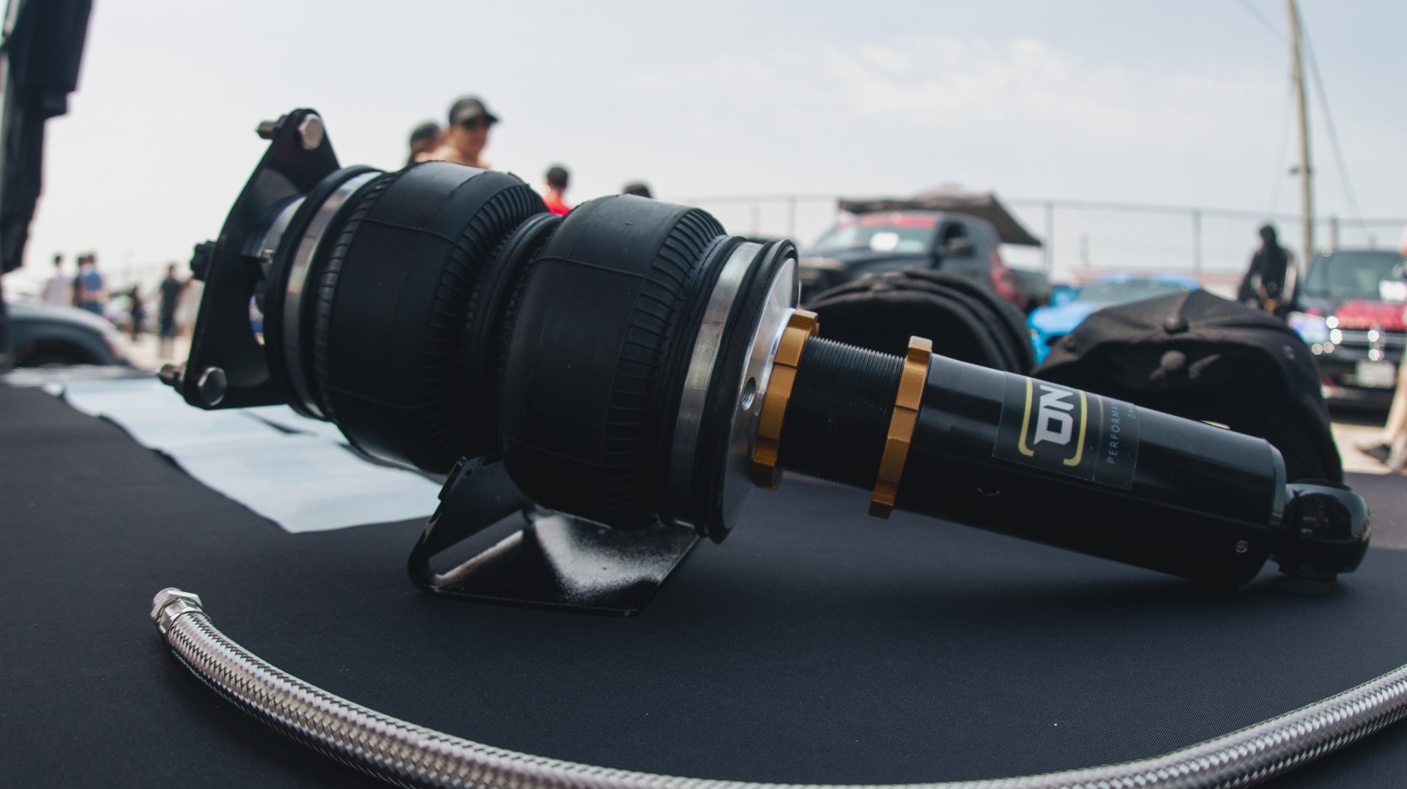 Image of this photo is a side shot taken of an ONAIR Performance Air Ride Strut. featuring a stainless steel braided line for the airstrut, the air bag is complimented with gold collar rings and adjustable camber plate at the top. behind the air strut are some onair hats