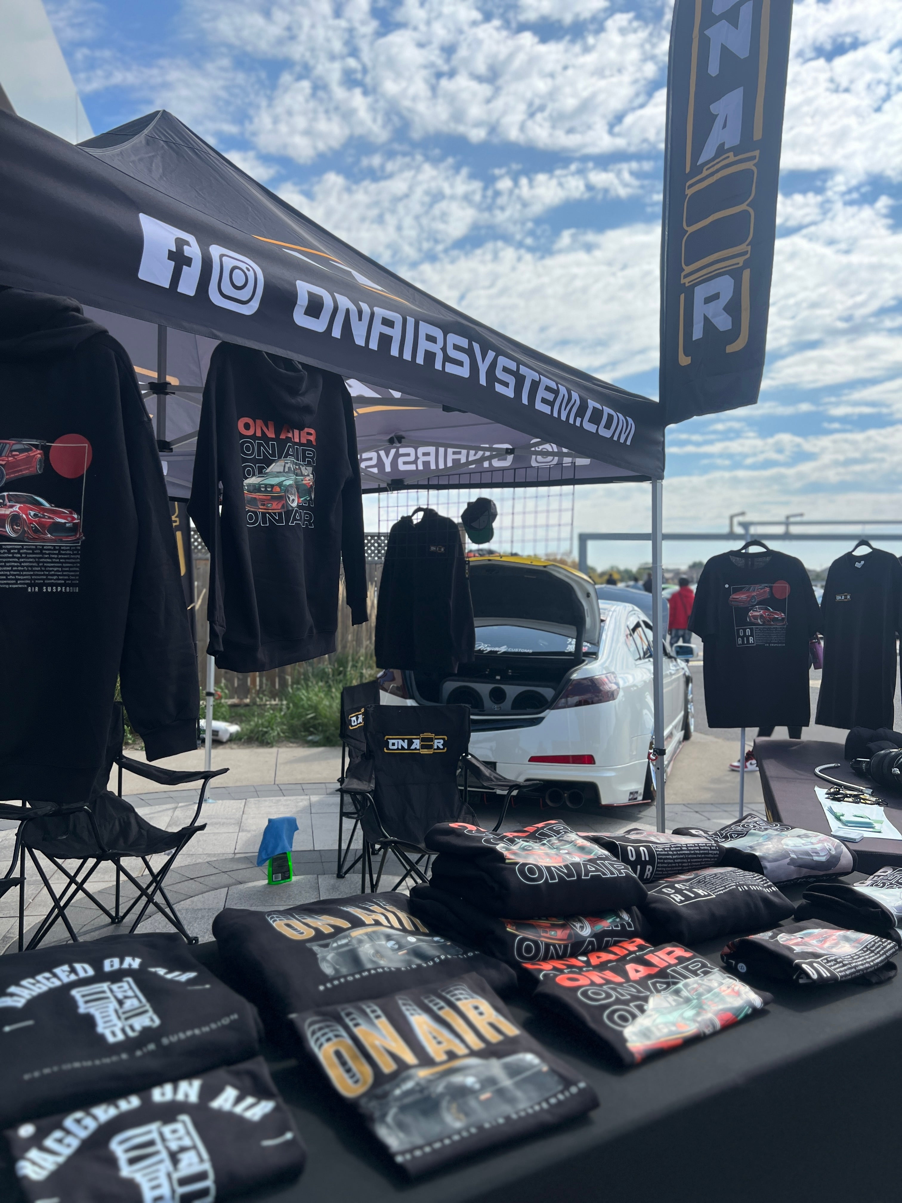 photo taken of onair vendor spot with onair merch featuring bagged on air hoodie and tshirts onair porsche onair bmw and a few other iconic desgins. there are flags in the shot and lots of branding